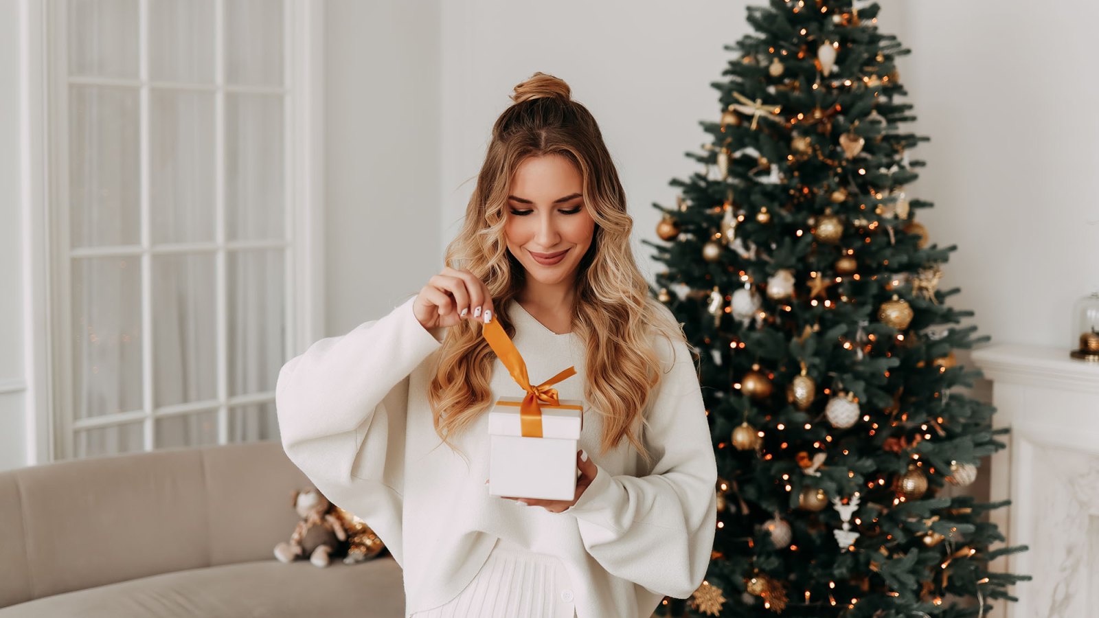 The concept of Christmas. Portrait of a smiling young woman with curly hair in a knitted sweater holding and opening a Christmas gift on the background of a Christmas tree in a bright interior on a holiday at home