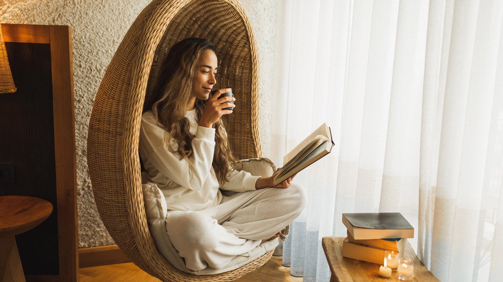 Woman relaxing at home in hanging ball chair, reading book and drinking hot herbal tea