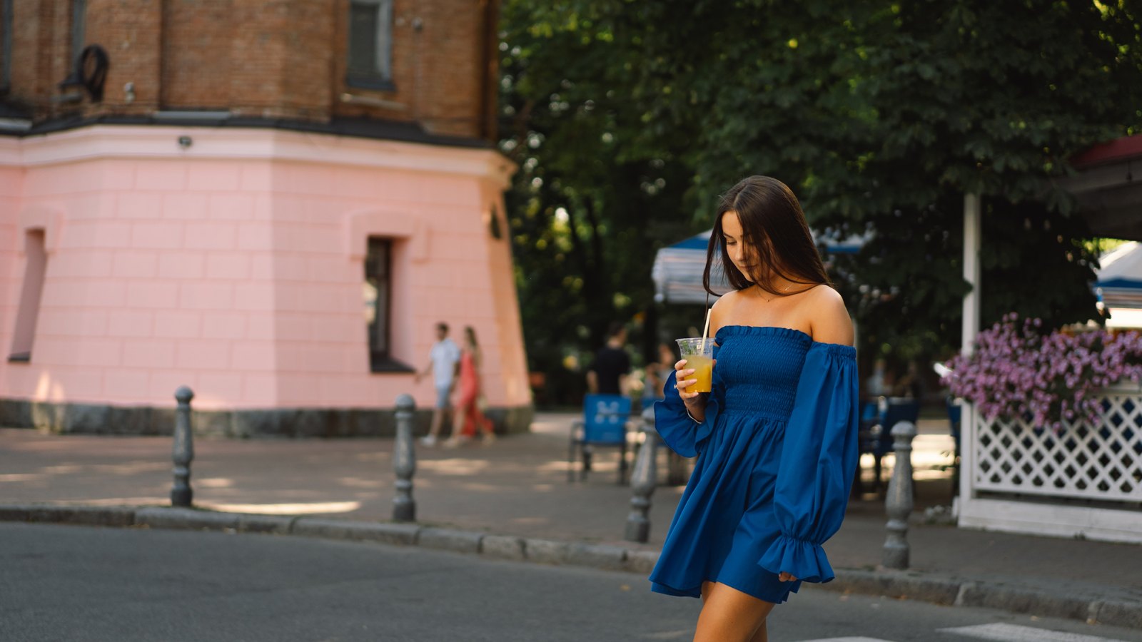 Portrait happy young woman wearing blue dress and drinking summer cocktail lemonade, standing on the street. Urban background.