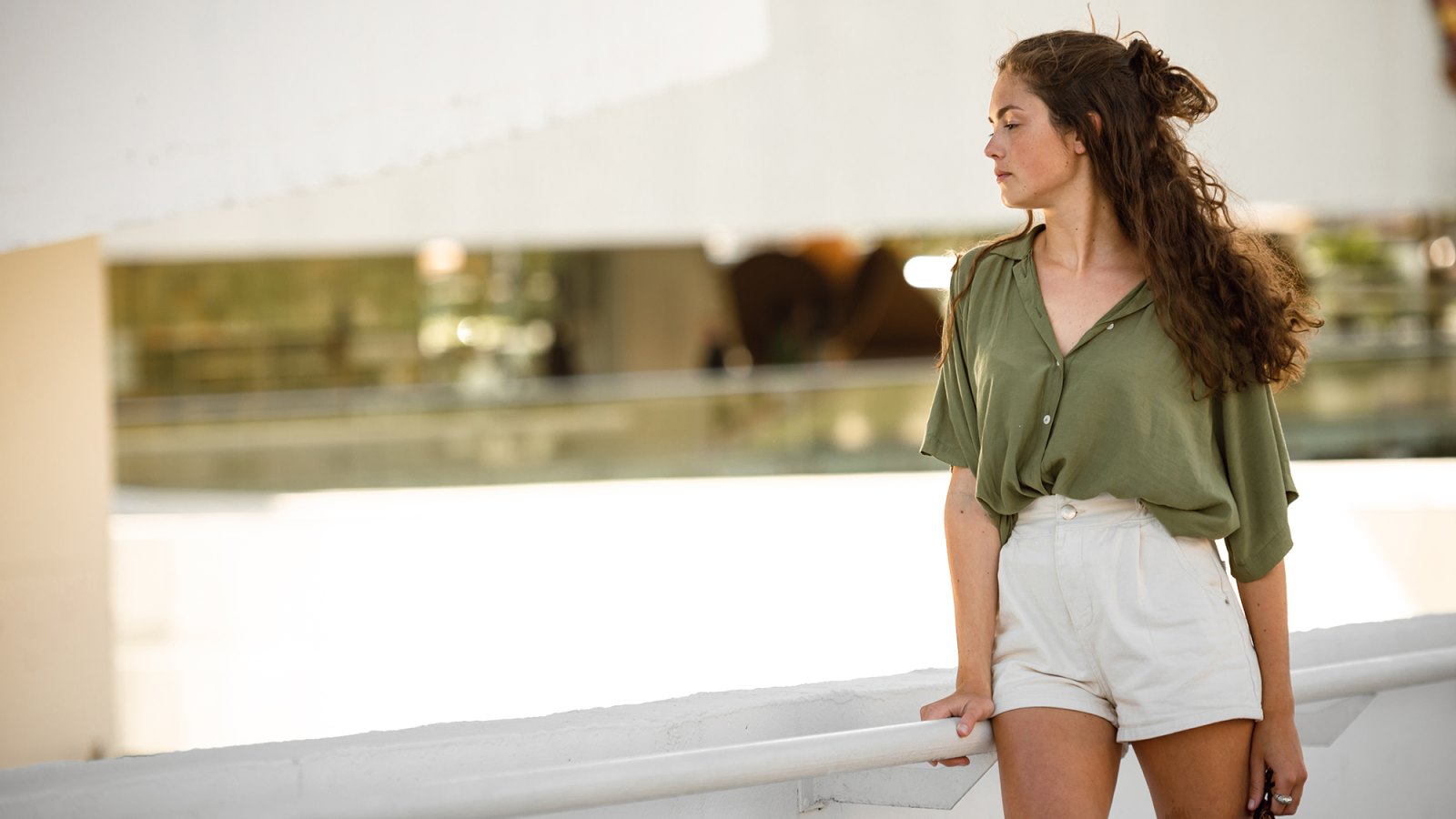 Portrait of a young adult woman, wearing a green t-shirt and a white shorts, leaning on a handrail and looking to her right.