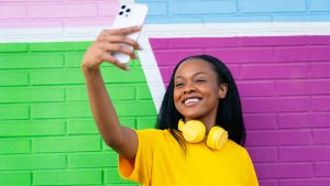 Brightly dressed woman with yellow headphones enjoys taking a selfie, with a vibrant urban wall behind.