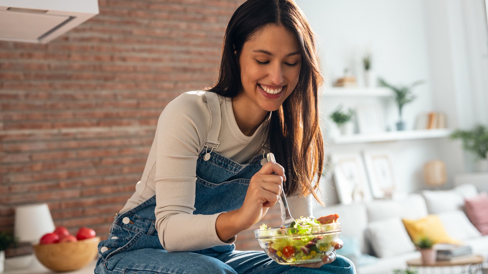 Shot of smiling woman eating healthy salad while sitting on the kitchen table at home.