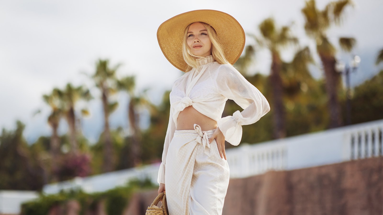 Stunning blonde woman in summer beach outfit relaxing outdoors against sea resort and palm trees on the background. A fashionable romantic young adult lady wearing a trendy vintage straw hat, white blouse, and skirt, standing at the sea coast