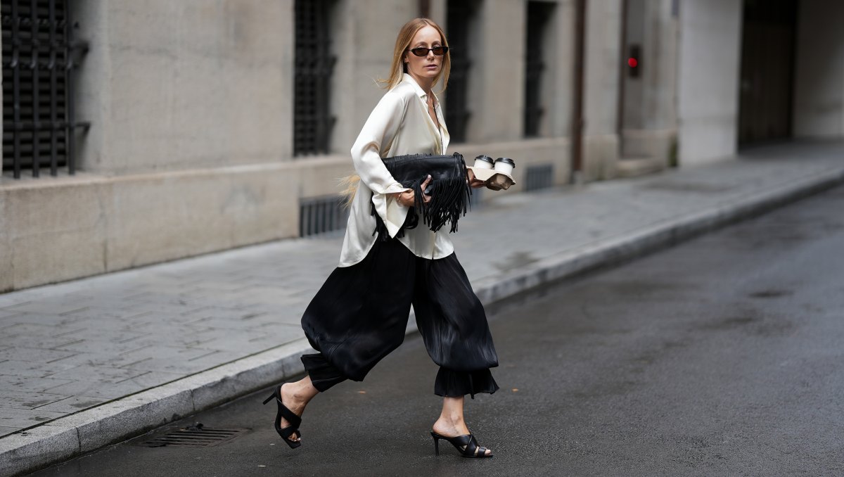 MUNICH, GERMANY - AUGUST 20: Anouk Delphine is seen wearing a white silk blouse from Lillysilk over a white bralette top from byAylinKönig; black balloon trousers from NA-KD; black leather heeled sandals from Zara; a black fringed leather shoulder bag from Zara; a floral necklace and a gold chain necklace from Cartier; gold rings from Wempe and Cartier; takeaway coffee cups; oval frame sunglasses with dark tinted lenses from Gucci; long blonde hair worn loose on August 20, 2026 in Munich, Germany. (Photo by Moritz Scholz/Getty Images)