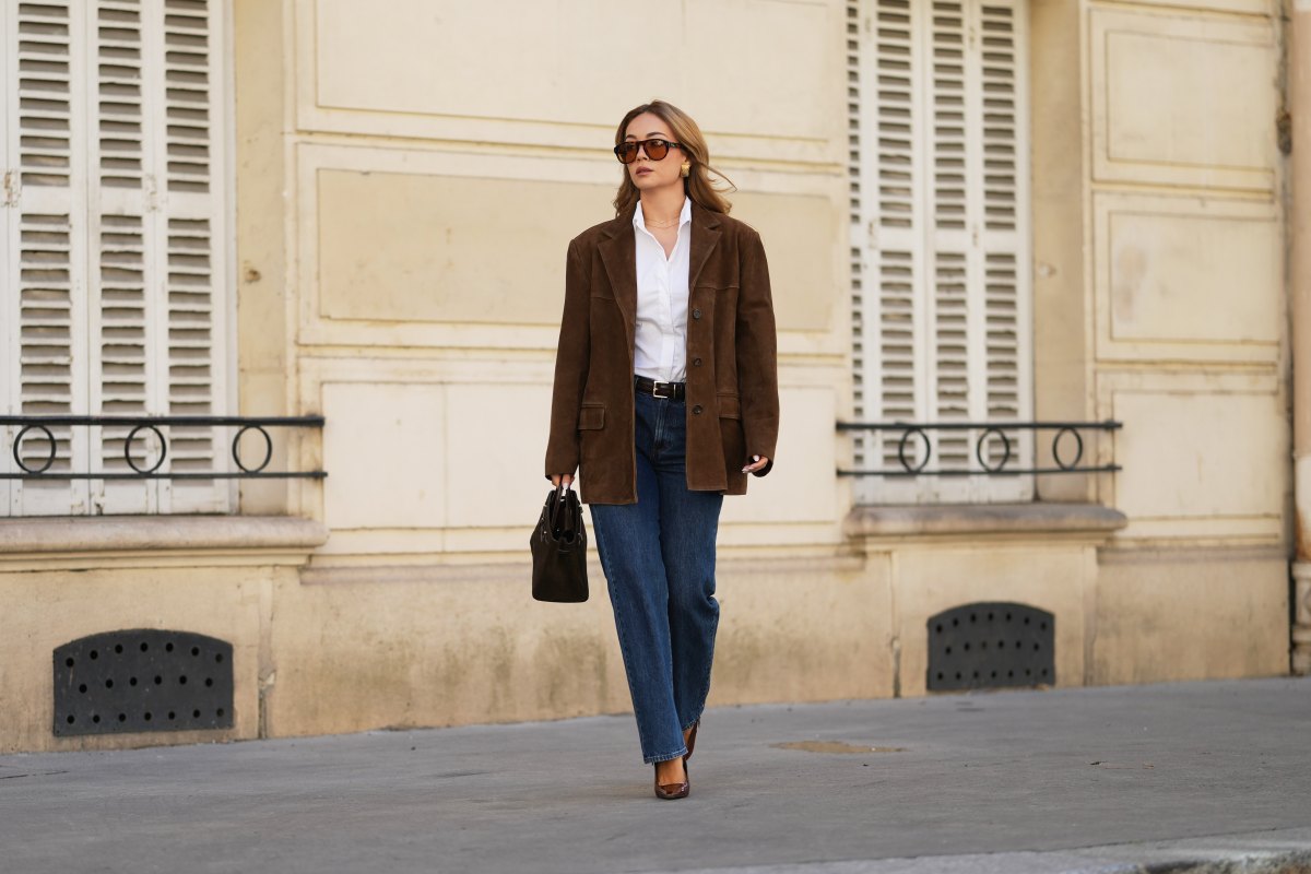 Merve Gorgoz wears brown long hair with center part, amber colored sunglasses with orange tinted lenses by Jimmy Fairly, golden square earrings, a brown suede oversize blazer jacket by Source Unknown, a white shirt, a leather belt, blue denim pants jeans by Mango, a dark brown khaki bag in suede by Demellier, brown pointed shiny patent leather shoes with high heels by YSL Saint Laurent, during a street style fashion photo session, on September 01, 2026 in Paris, France.